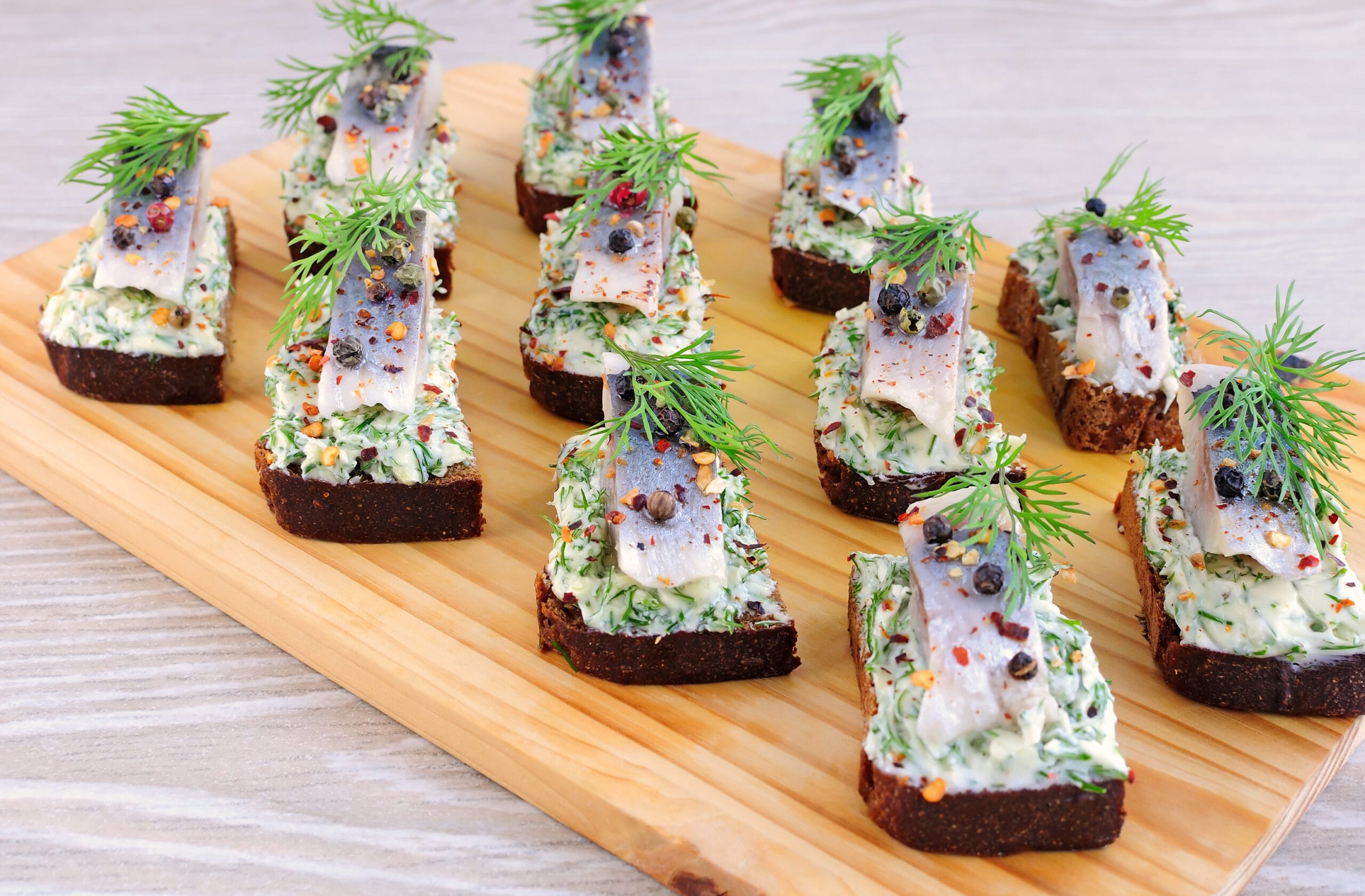 Wooden board of dark rye canapes topped with herring, herbed cream cheese, dill, and peppercorns